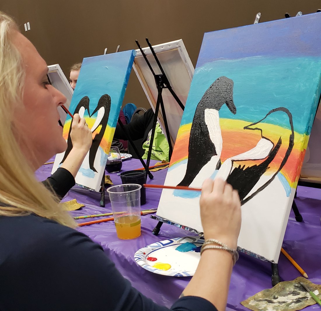 Meredith in profile at a paint-and-sip class, brush in hand, working on a canvas of two black-and-white penguins against a rainbow sunset.